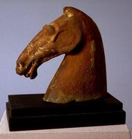 A gray earthenware head from a horse sculpture; its strong muscular neck holding its narrow head high. It is vividly sculpted to show the musculature of the horse's face with flaring nostrils and an open mouth showing its tongue. It has deep set and bulging eyes, ears pointing forward and a flowing mane. It is covered in red mineral pigment. 
