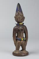 Standing female figure on a round base with hands at the sides. Around the waist and neck are strings of multi-colored beads. On each cheek of the face there are three incised marks and the pupils of the eyes are metal. The hair is in the shape of a rounded comb, with horizontal grooves coated in blue pigment. 