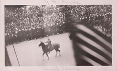 A view from above of a military parade. A man in uniform on a horse is clearly visible in the center of the image and a flag, a portion of which is striped, obscures figures behind the man on the horse. The parade is bordered by many people wearing dark clothing and white hats.&nbsp;