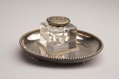 Inkwell made with crystal and glass has a cube shape body and a metal lid. It is placed on a silver round tray with beaded edge surrounding the tray edge.