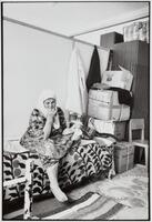 An older woman seated on a bed covered in blankets. Her head is covered in a scarf and there are boxes piled in the corner.