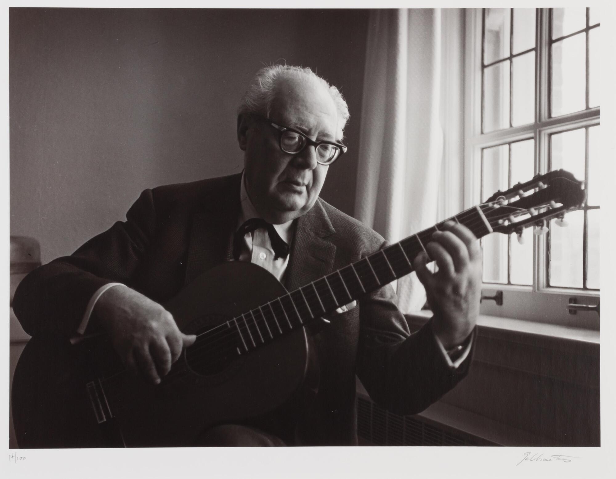 Older man playing guitar next to a window. He is wearing glasses, a button down shirt and a jacket.
