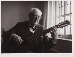 Older man playing guitar next to a window. He is wearing glasses, a button down shirt and a jacket.