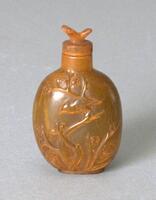 An orange rhinoceros horn snuff bottle with birds and tree branches carved on the surface in raised relief. On the top is a stopper with a bird figurine on the top.
