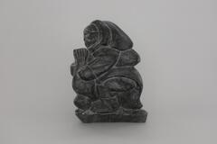 A carving in black stone of a man wearing a parka and boots, holding onto a seal with both hands.&nbsp;
