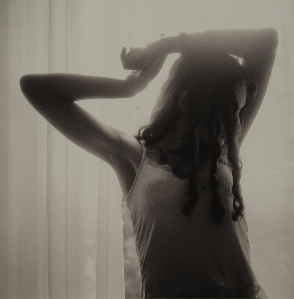 This is a photograph of a young girl silhouetted in front of a bright window with her arms raised above her head. Her left arm rests on her head, while she clasps her wrist with her right hand.
