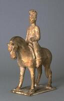 An earthenware figure of a horse and rider. The horse is standing on all fours on top of a rectangular slab base. It has a trimmed mane and long flowing tail, the male rider sitting tall and wearing a long coat, trousers, a tall cap, and boots. The rider has one hand in a position to hold reins. The figure is covered in a straw-colored glaze.