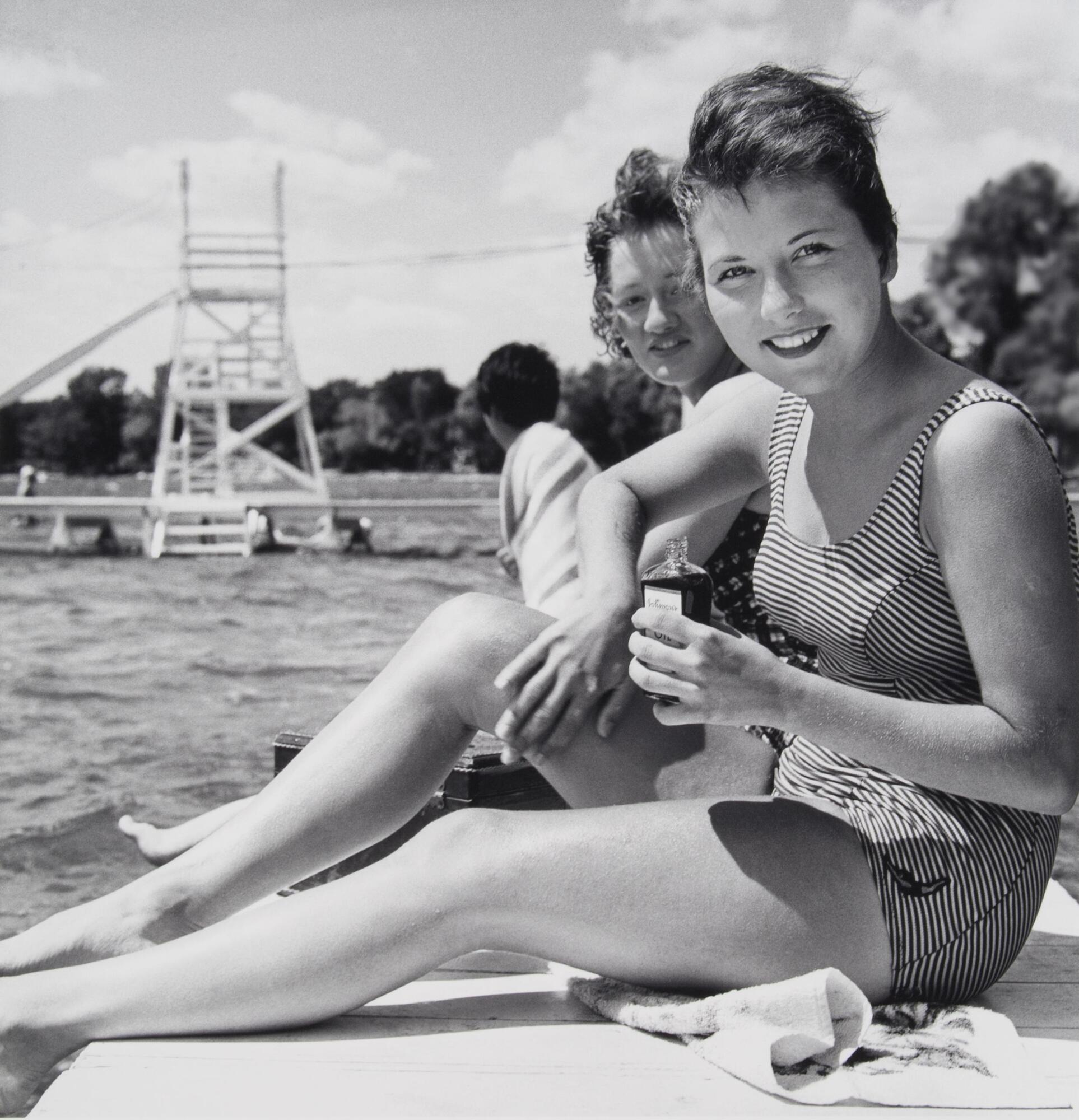 Two women seated on dock in bathing suits, looking at the camera. The woman in the front is holding a small clear bottle.