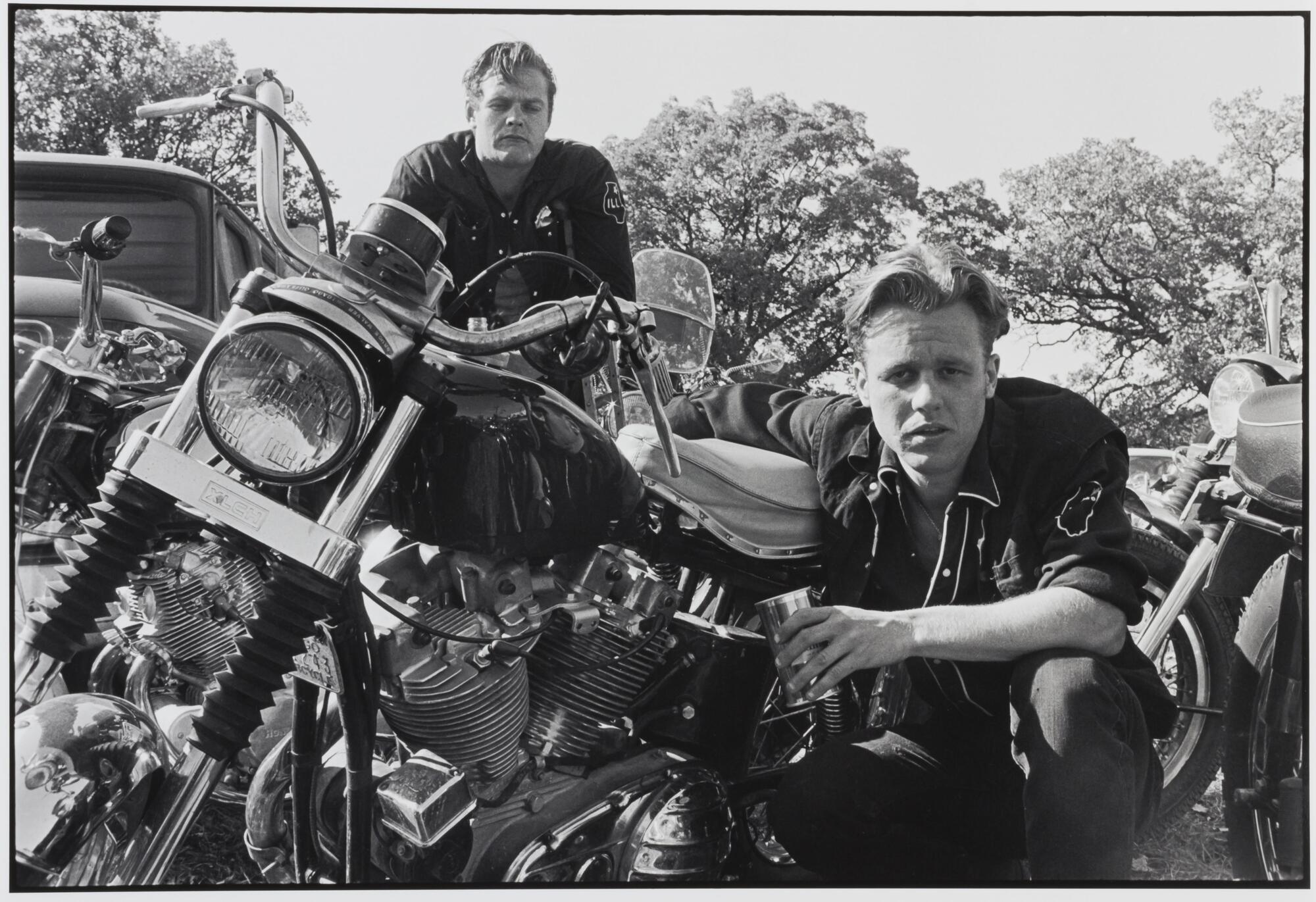 A photograph of two men with motorcycles. One man sits atop a motorcycle to the left. The second man crouches beside another bike, his right arm resting on the seat, a cup in his left hand. Both men look into the camera lens.