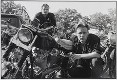 A photograph of two men with motorcycles. One man sits atop a motorcycle to the left. The second man crouches beside another bike, his right arm resting on the seat, a cup in his left hand. Both men look into the camera lens.