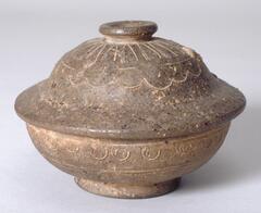 Stoneware jar with natural ash glaze and bell-shaped lid. Along the widest horizontal stretch of the base is an incised circle-and-dot design. A variation on this pattern also loops around the lid, topped by a line marking a ring along the surface of the lid, and incised lines extending outward from a button-shaped knob.<br />
<br />
This is a dark gray, high-fired stoneware lidded bowl with a stamped design. The lid features a button-shaped knob at its center. Thinly incised lines encircle the upper part of the lid, dividing its surface into two segments. The inner segment is decorated with a triangular line design, whereas the outer segment is decorated with a design consisting of semicircles with dots inside, which border the inner segment. Sets of two thinly incised horizontal lines run around the section of the bowl immediately below the rim and the lower middle part of the body. A double semicircular design decorates the segment between these two sets of lines. The foot is short and thick and has a fo
