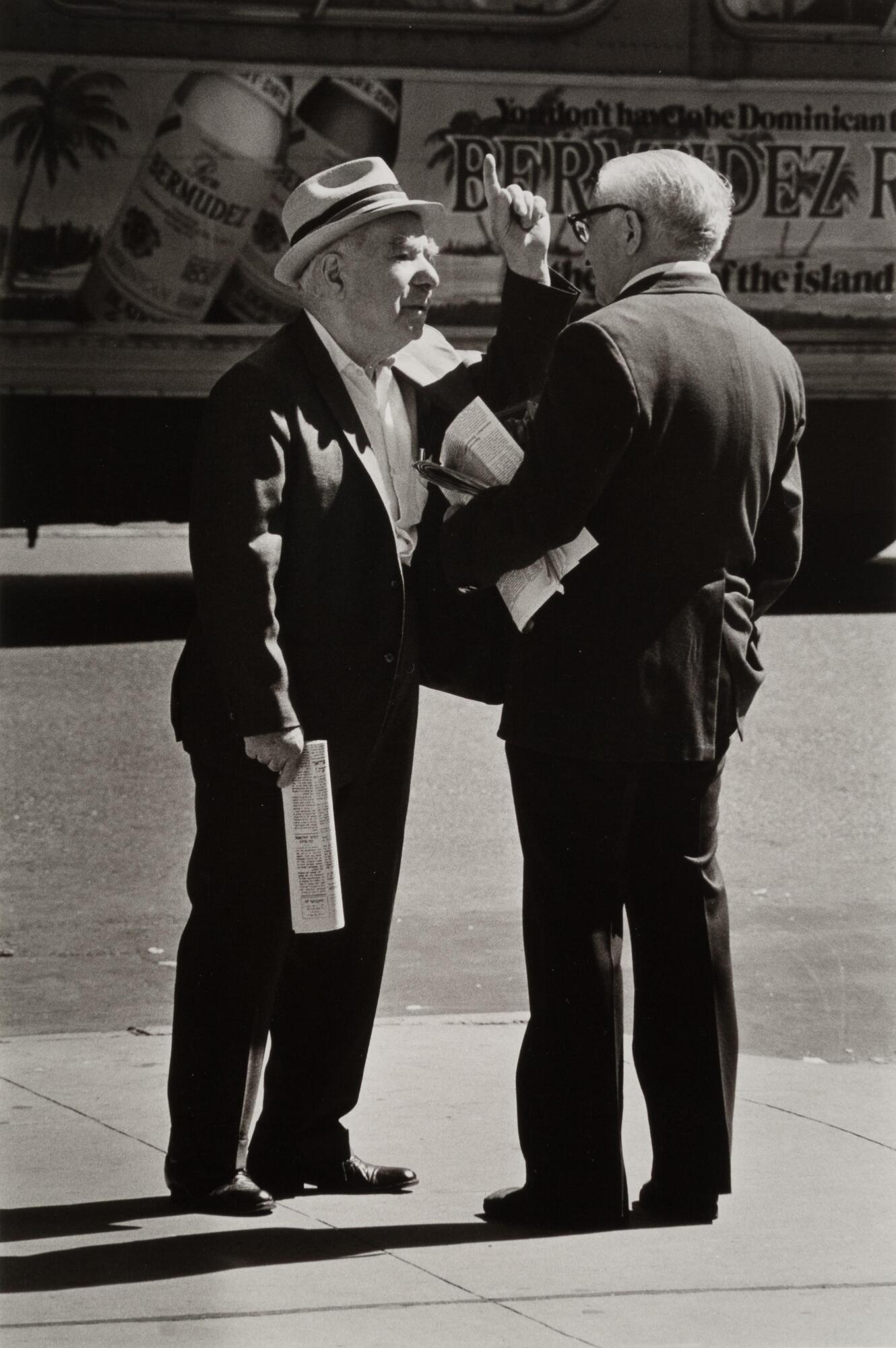 Two men in suits on a sidewalk, both holding newspapers, one his holding a hand up, they are in the middle of a conversation.
