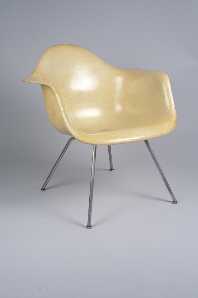 Pale, yellowed plastic armchair, consisting of a single "shell" of compound curves, slightly tilted back on a four-legged metal base.