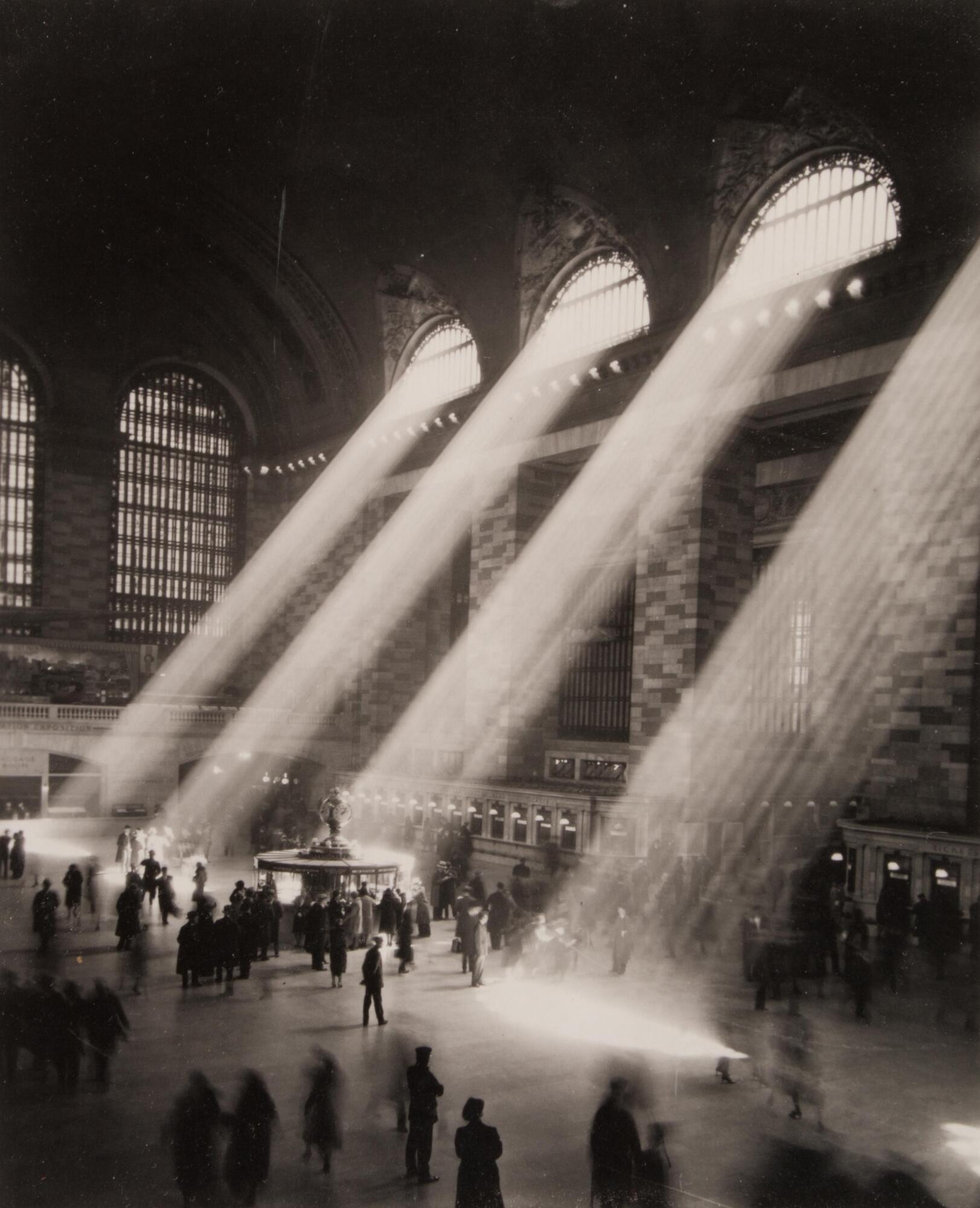 A black and white photograph of a cavernous building. Beams of light stream in through the windows, revealing a smoky atmosphere as people move about the main concourse below.
