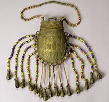 An oval-shaped brass purse with loops around the outside edge. Attached to each loop is a string of beads or leather ending in a brass crotal bell. The body of the purse has raised ovals with a spiral pattern inside. The lid has a brass handle and a longer beaded strap. 