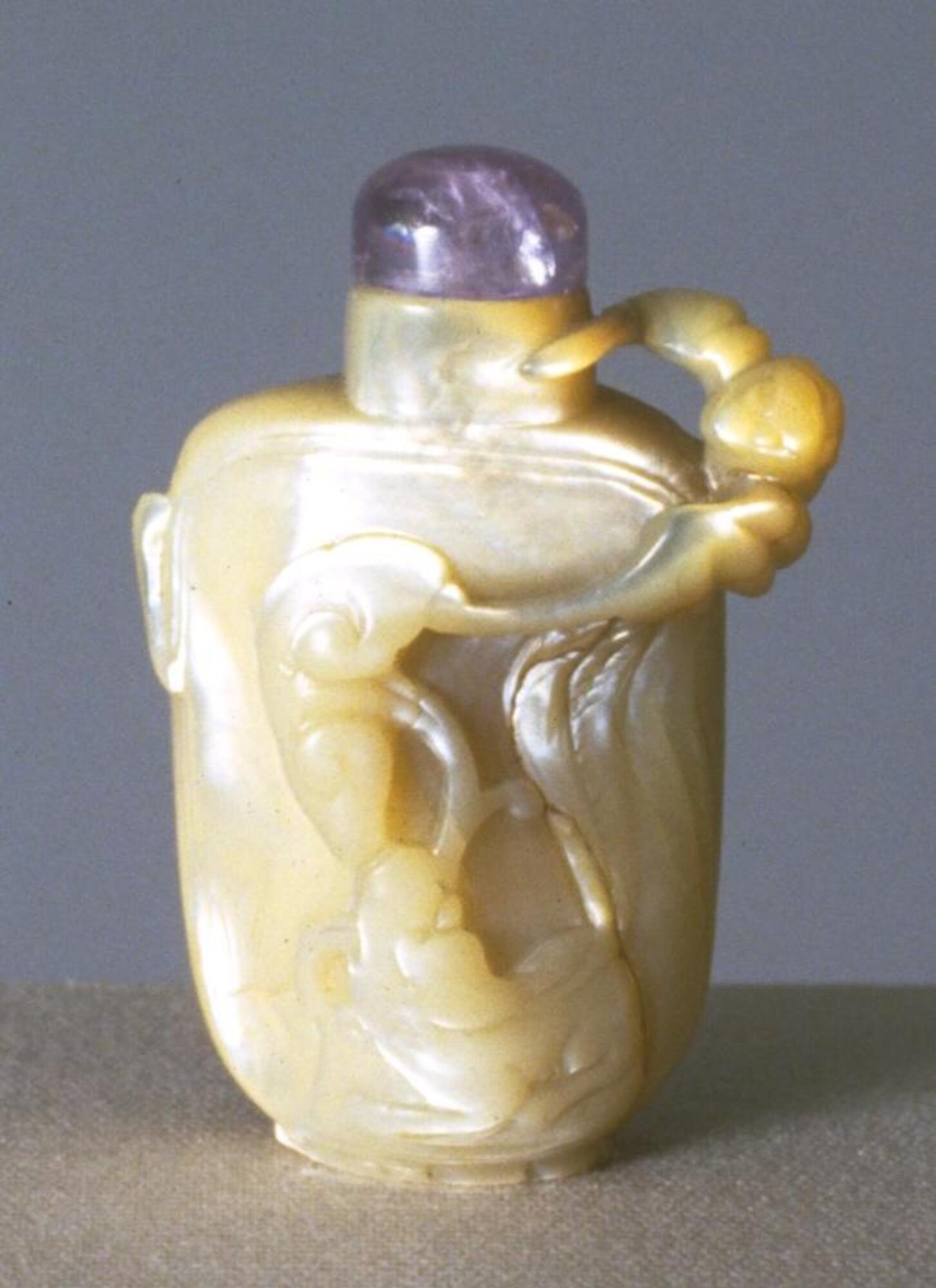 A mother of pearl snuff bottle carved with organic designs. On top of the snuff bottle is an amethyst stopper.