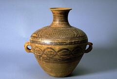 A light reddish-buff earthenware&nbsp;<em>guan (</em>罐) jar with a wide globular upper body and conical lower body on a flat base, and a tall narrow neck with an everted rim. There are two diametrically opposed lug handles at the waist. The upper half of the body is painted with black and red pigments to depict four whirls of concentric circles. Each contains a roundel made of joined circles, which also create a cross through the center. The four circular motifs are confined between solid band borders, with a garland border below. Around the neck are bold thick black chevrons.
