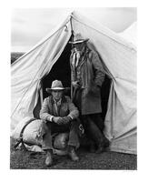 A black and white phot of two men dressed in a Western style with a tent. One man is standing in front of the tent, while the other is sitting on a rolled up canvs in front of the tent.