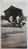 Two prop human skeletons from the shoulders up, dressed in shirts and hats and placed under a black umbrella. The skeletons appear to be sitting on a wooden pier with water visible in the background.