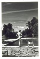A photograph of a nude woman standing and looking over a pool with palm trees in the background and a director's chair to her right in the foreground.