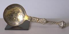 This silver ball spoon consists of a round bowl joined to a stem composed of a flattened section and a twist stem that terminates in an ornamental ball knop. An incised six-petal flower surrounded by bands of geometric ornament decorates the interior of the bowl. The flattened section of the stem is adorned with vegetal ornament.