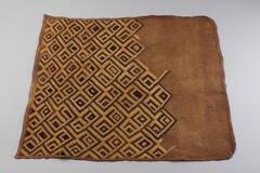 Rectangled panel with hemmed edge consisting of two squared and two rounded edges. There are tan and brown trangular patterns partially covering the panel consisting of uneven chevrons and interlocking rectiliniar designs.