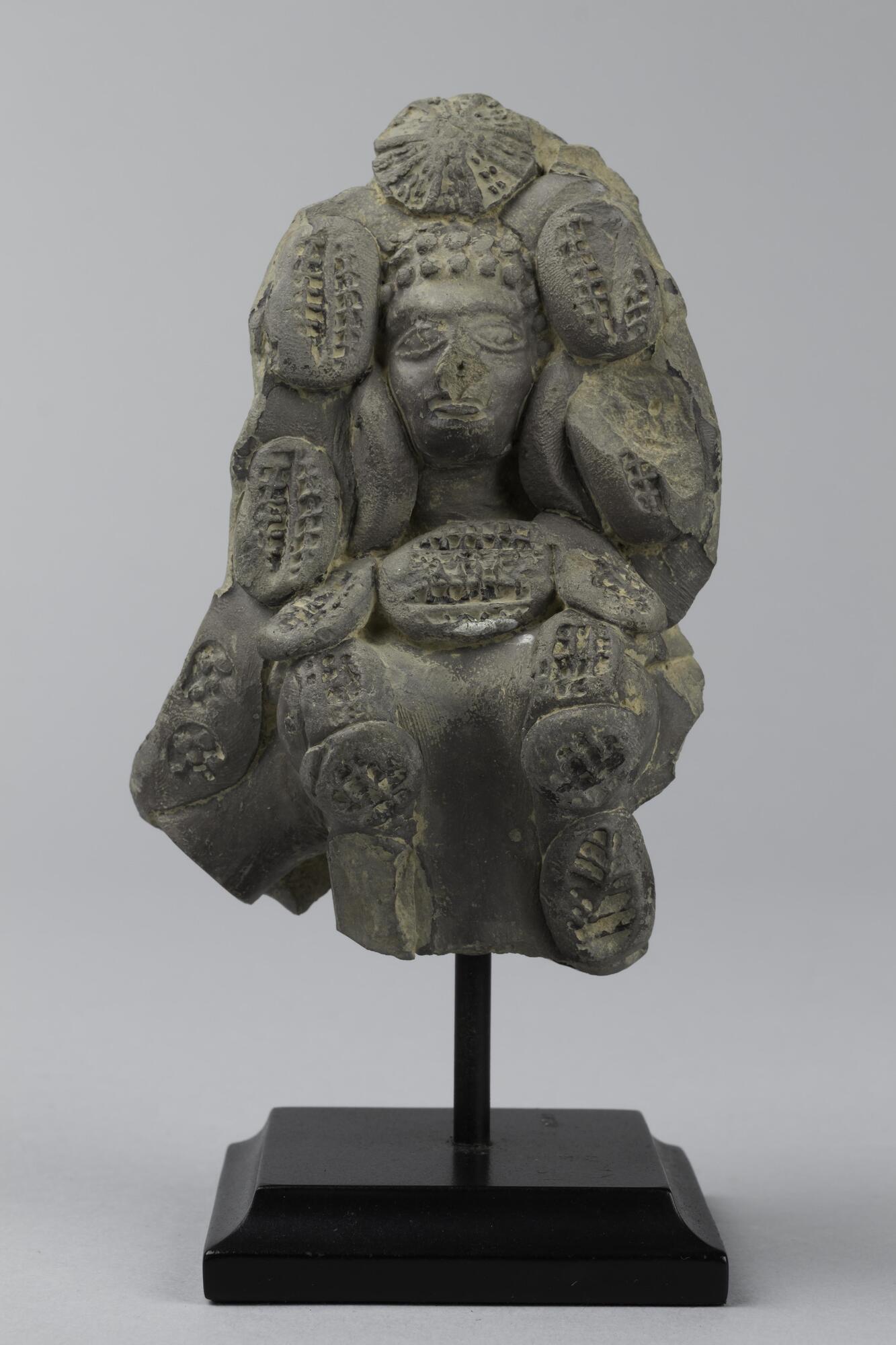 This black terracotta is broken at the waste and depicts a simple body decorated with globs of clay decorated with punch-marked decorations.  The face is modeled a bit more realistically with wide-open eyes and a full mouth.  The front of her hair is in a series of rounded ball-like curls and a circular starburst decoration is at the top.  The other oval globs frame the face of the goddess and cascade down what is left of her body.<br />