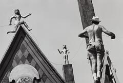 A man attaches wires to a wooden pole in the right foreground; humanoid sculptures adorn a pointed roof and chimney on the left. 