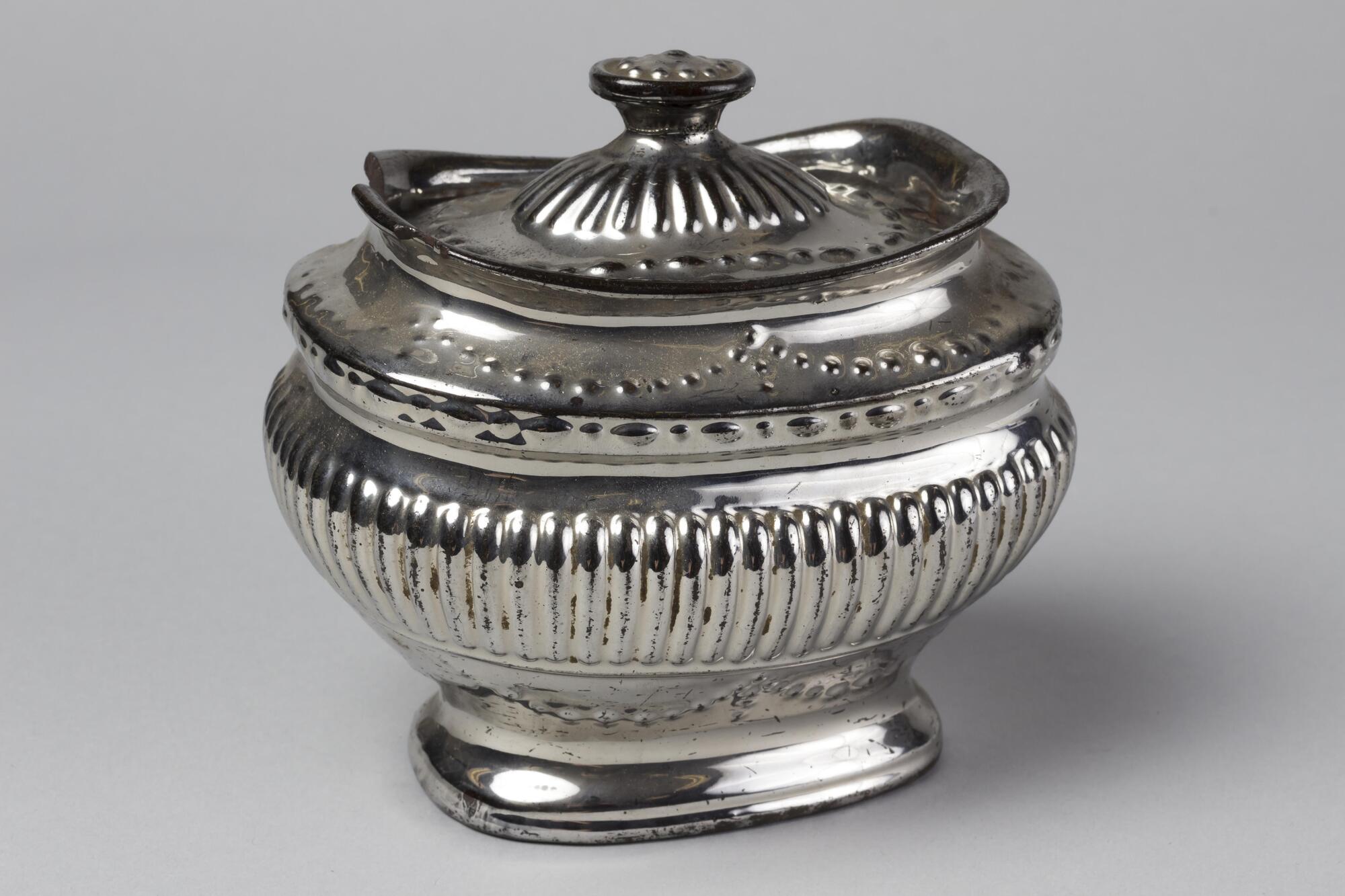 Silver jar with a scalloped dot decoration and vertical lines. The top swoops up on the sides.