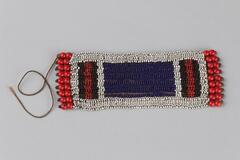 Beaded jewelry with a rectangular piece in white with blue rectangular center piece and red and black striped sections on either end. Large red beaded fringe on two ends. One long thread coming off end.
