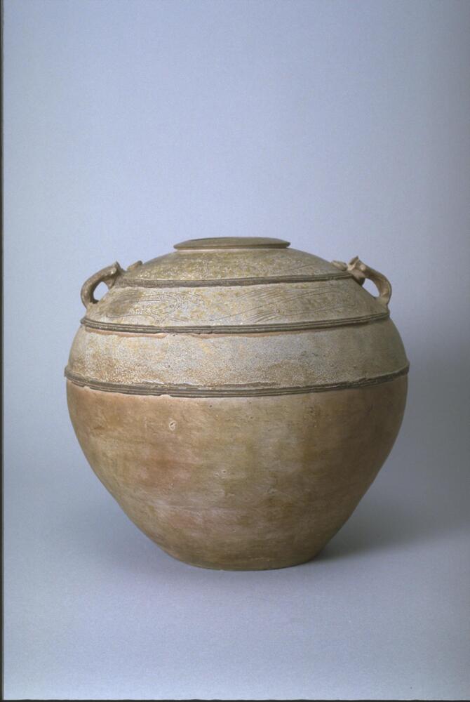 A brown stoneware jar (罐 <em>guan)</em> with a bulbous body, narrow mouth with wide flattened rim, on a flat base, with appliqué bowstring bands around the body and rim, alternating with incised scroll work. There are two animal mask lug handles on opposing shoulders. The upper half covered in a gray-green ash glaze.