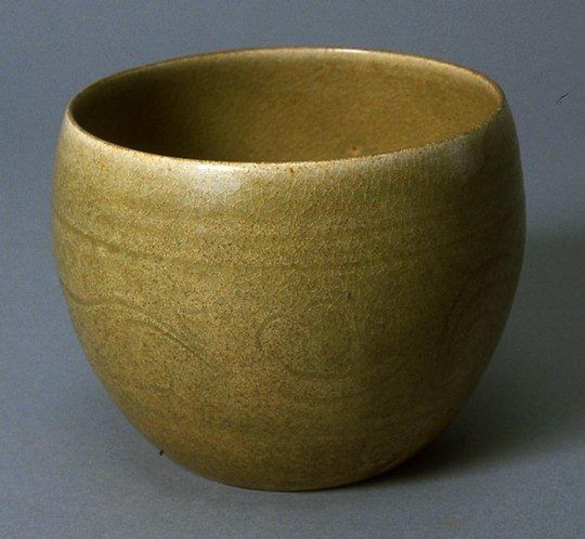 A finely potted stoneware beaker with a rounded bottom and a lightly incised, freely drawn leaf scroll spreading across the widest part of the body, flanked by incised double horizontal lines, all covered evenly with an yellow green glaze. Four spur markes inside.