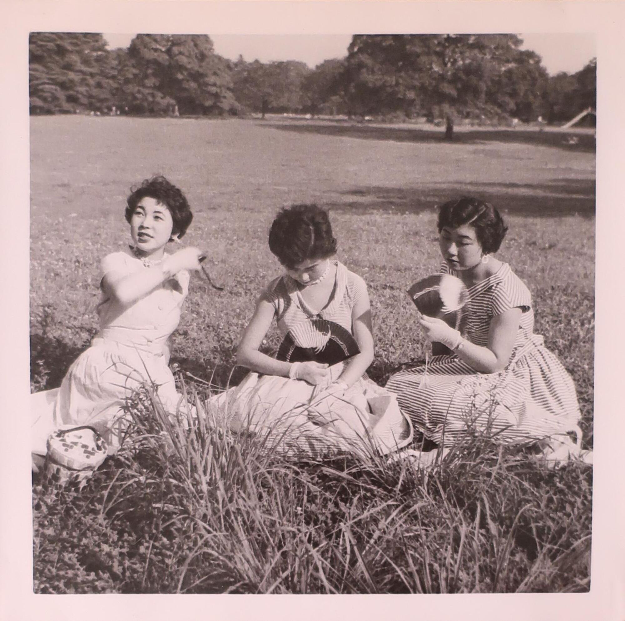 Three young women with short dark hair sitting in a field. The women on the right and in the middle hold fans in front of them. The woman on the left looks up towards the left side of the image and has her mouth slightly open as though speaking.