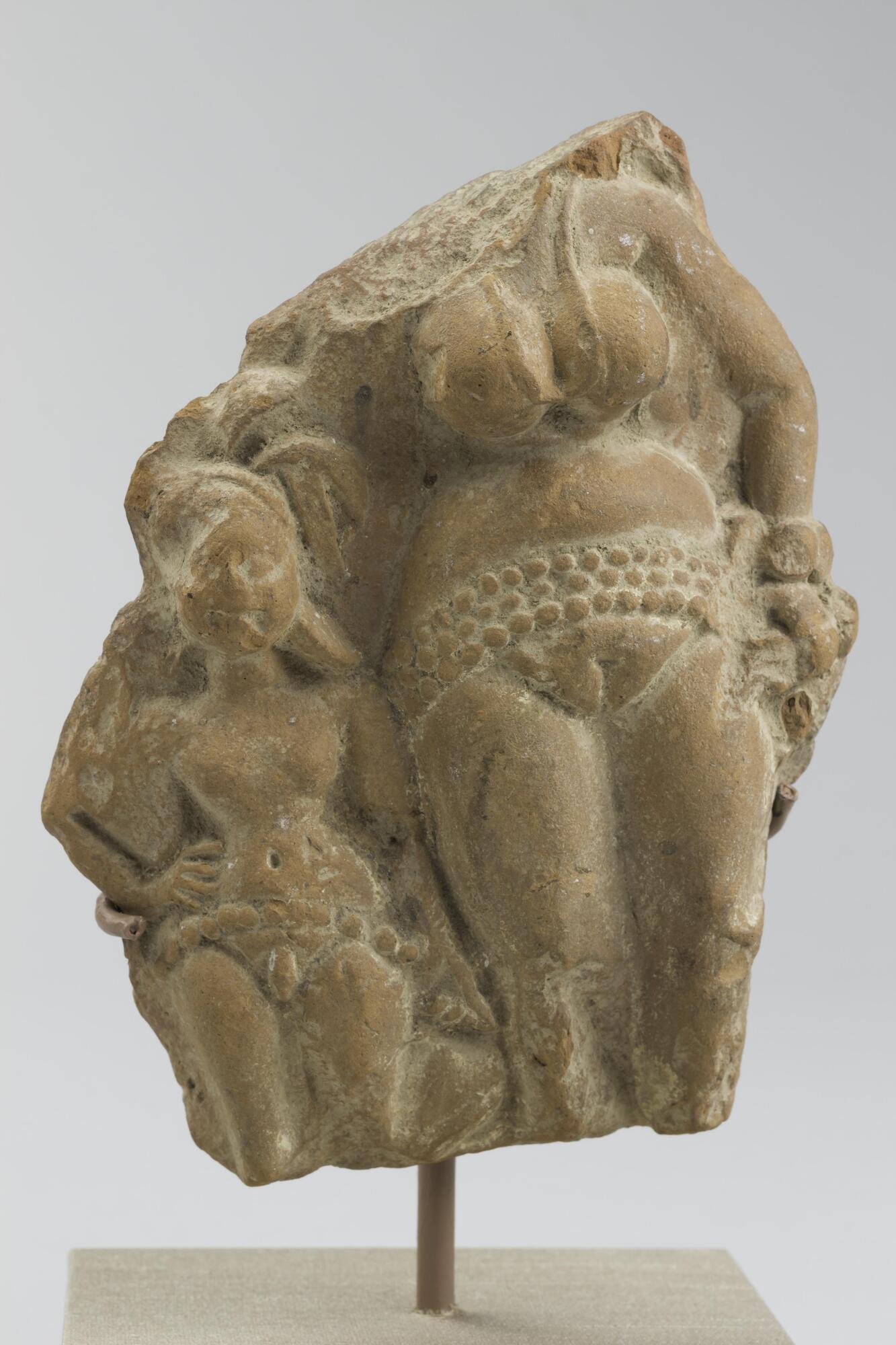 This broken fragment depicts a now headless goddess or yakshi standing with a much smaller male attendant.  She wears a necklace, bracelets and a girdle consisting of three rows of small round shapes.  She stands with her left hand at her waist and the right hand next to the head of her attendant.  He wears a turban and large earrings and a belt of a single line of round shapes.  They both wear diaphanous lower garments that make them appear nude with both of them with their genitals exposed.  The fragment is broken above the male figure’s knees and below the yakshi’s knees.  <br />