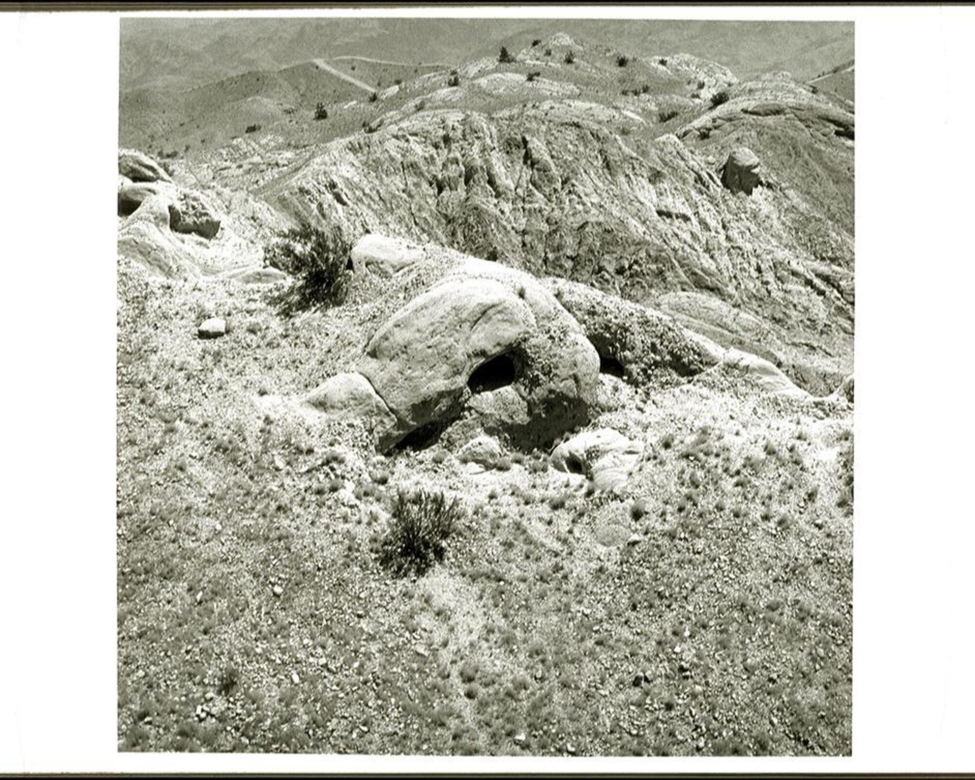 This is a black and white photograph depicting a rocky hillside in a barren landscape. There are craggy peaks in the background and some scrub type vegetation. The viewpoint is from above looking down.