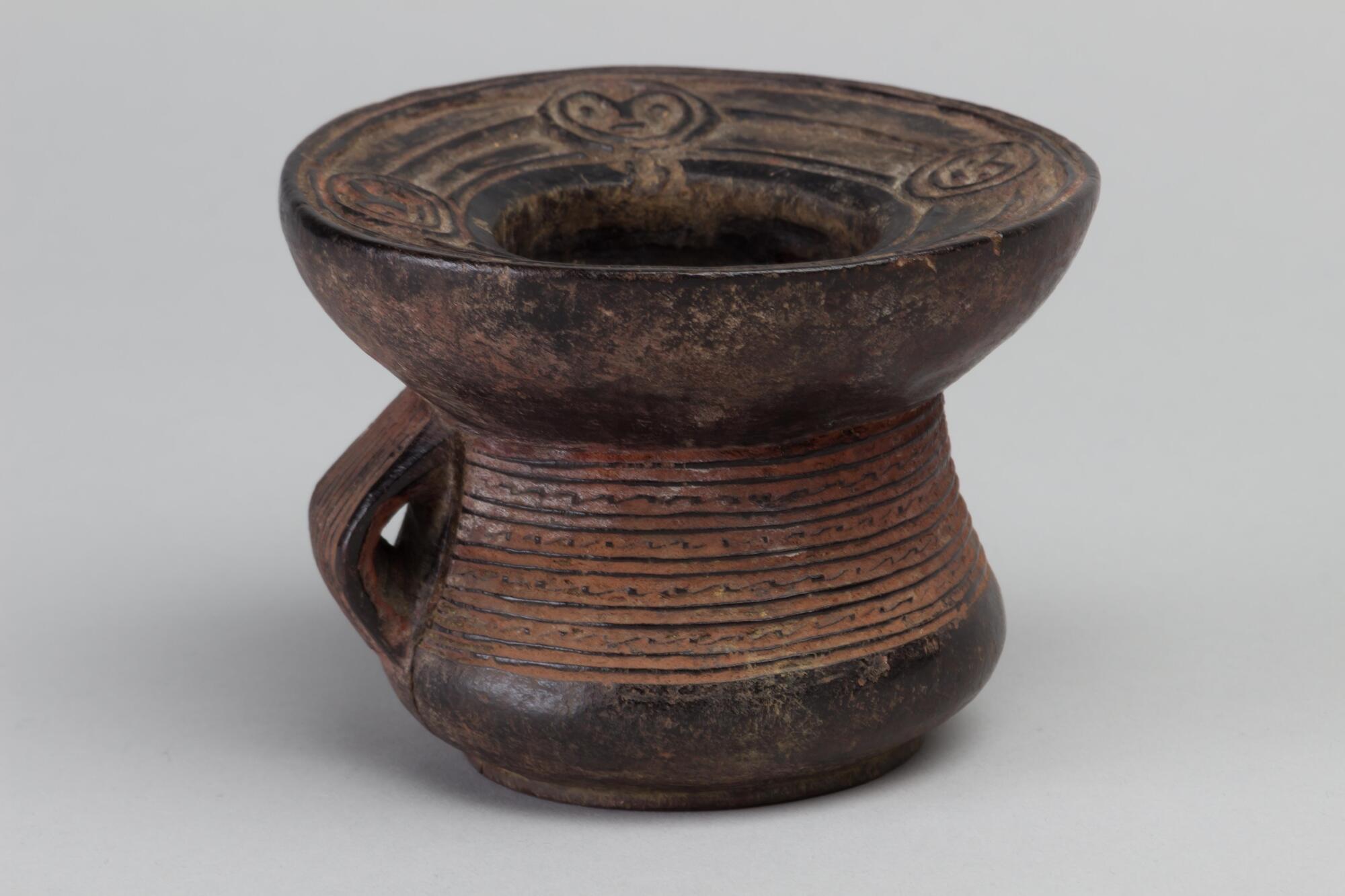 Cylindrical vessel with handle and a slightly buldging base. The rim is extended with faces designed on the inside of the rim's lip. There are linear design patterns wrapped around the cylinder portion of the vessel. 