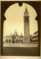 This photograph depicts a view of an empty piazza and a cathedral and tower at the far end. The image is framed by the shadowed form of an arched doorway.