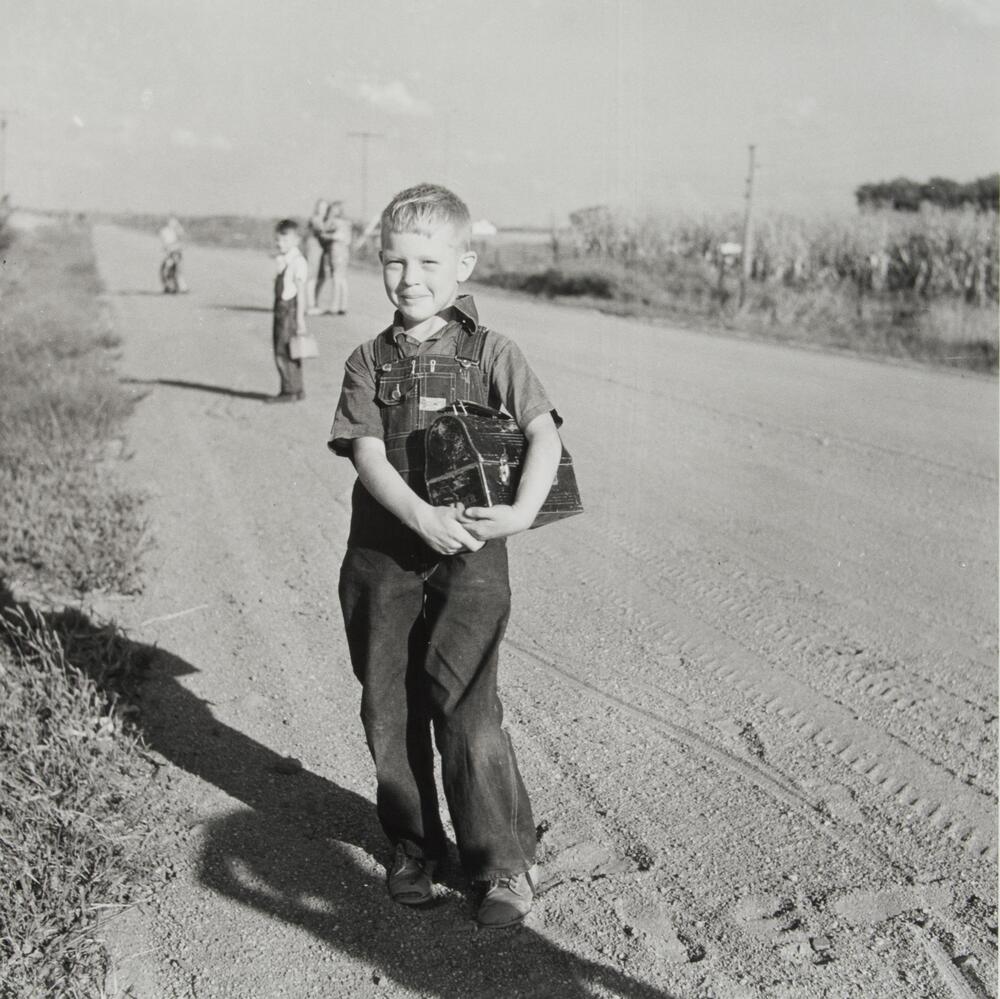 A boy on the side of a dirt road holding a metal lunch box in his hands. He is smiling and wearing overalls. There are other children in the distance.