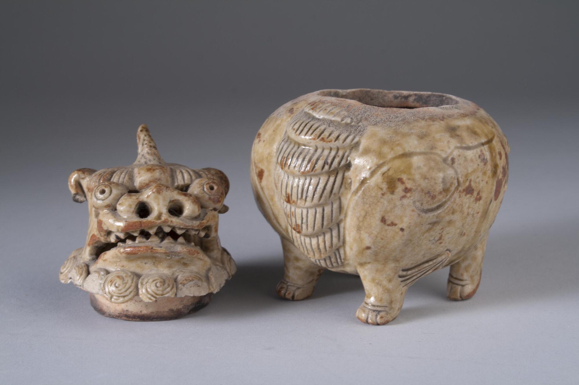 A container with an animal (dragon) shaped head as a lid; four paws at the bottom of the container. Glazed stoneware, sculpted with teeth incisions. Tan with red spots of coloring, partiallyy covered with olive colored gloss over a high-fire red clay. Small swirl design on sides and front area has carving to indicate the front/chest of the animal, sculpted with teeth and incisions.