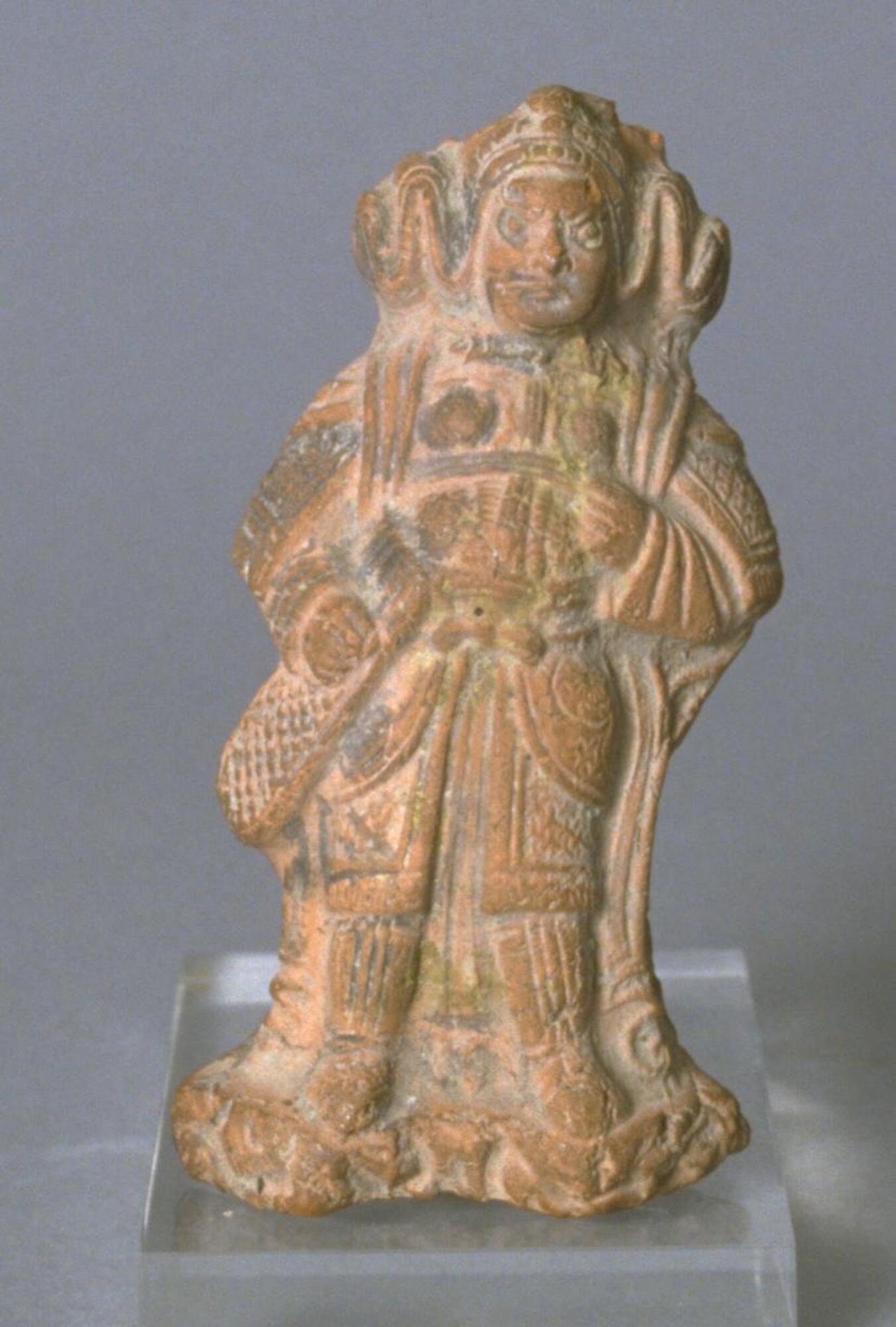 A red earthenware standing figure of Tianwang—Heavenly King, a Buddhist deity—wearing Tang dynasty styled armor including a helmet with flowing ribbons, elbow-length gauntlets, a cuirass with plaques, and taces, knee length greaves, and boots. The figure is standing on a rock-styled base, unglazed with traces of mineral pigment. 