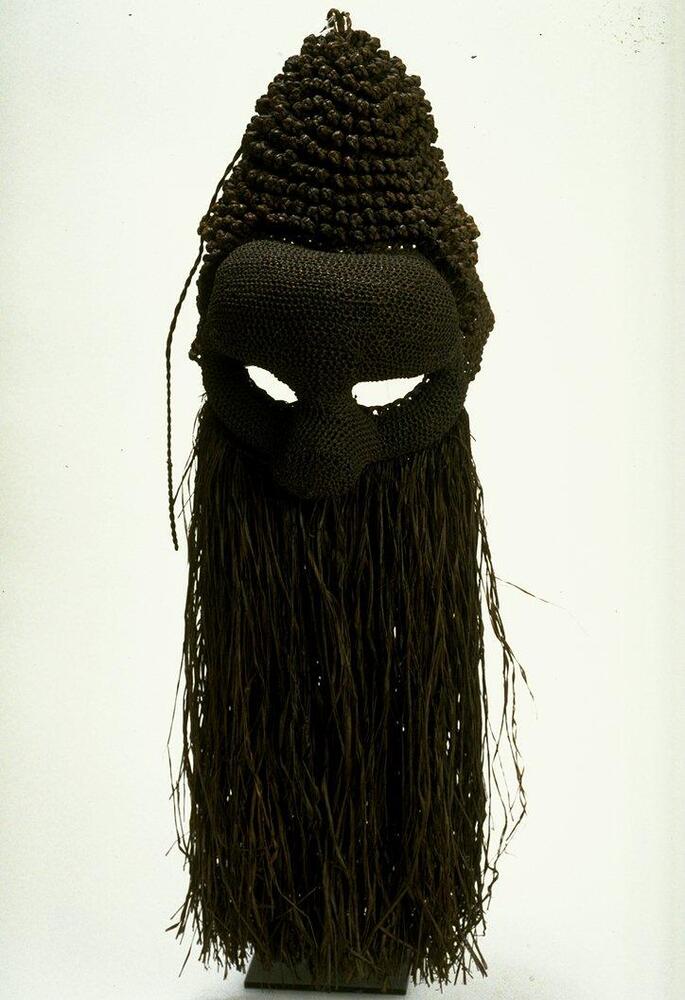 Mask is made entirely of blackish-brown dyed and molded raffia fiber; face has bulging forehead, deeo set narrow eyes, bulbous nose, and raffia “beard.” Top of head has cone-like crest of small fiber knots.<br />
