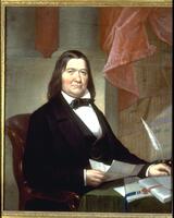 Portrait of man with brown, shoulder-length hair wearing black jacket and neck tie, seated at a desk holding unfolded letter. Set against a backdrop of whilte columns, red/pink drapery, and a bookshelf. Larson 2/5/18<br />
<br />
&nbsp;