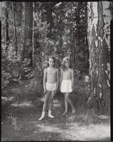Two girls, both wearing white bottoms only, one is also wearing socks, standing amongst trees with the sunshine pouring in.