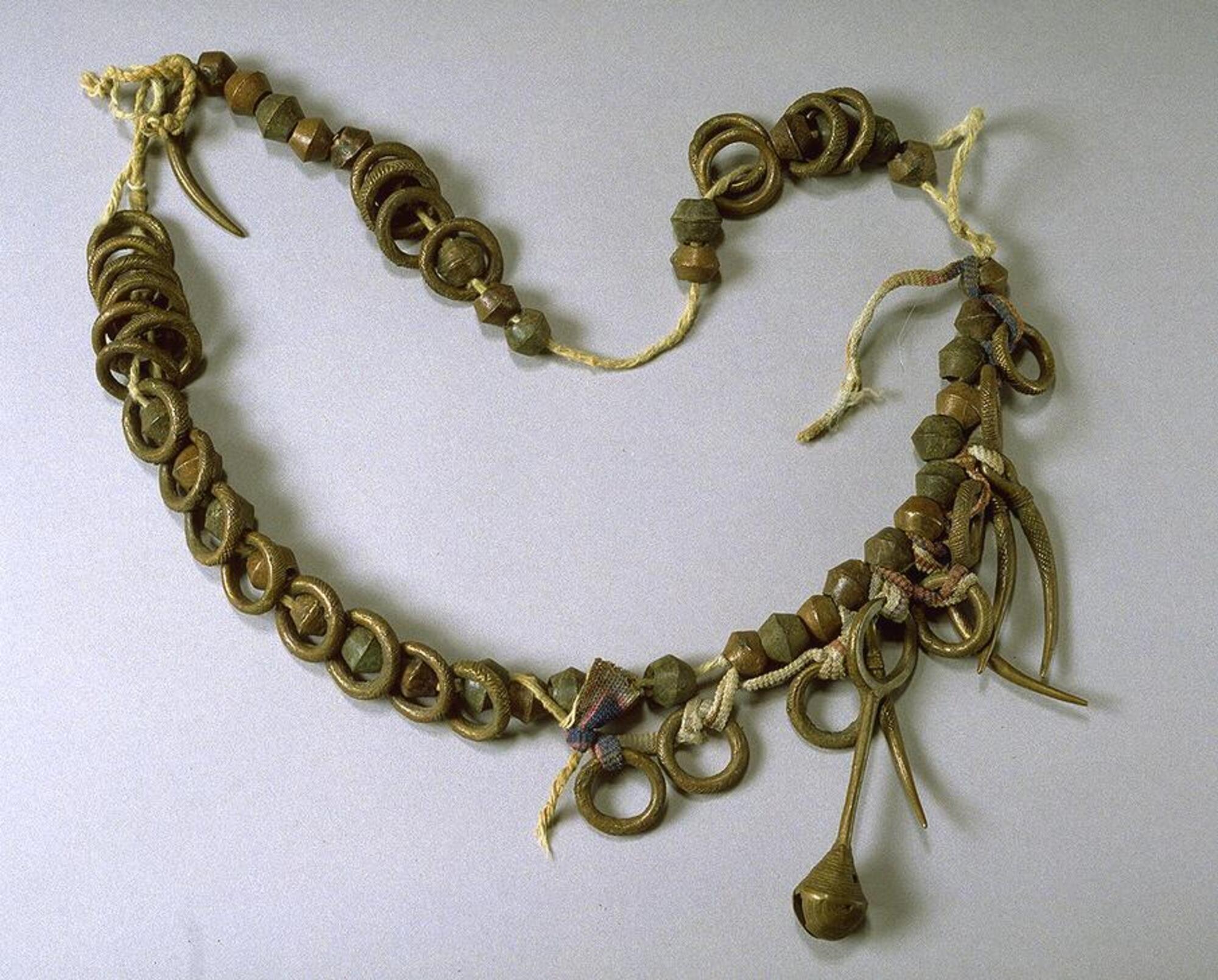 Belt with large, bicone brass beads and large brass rings threaded on a thick string. Some of the brass rings have incised grooves. There is one crotal bell pendant and five curved pendants. 