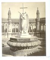 This photograph depicts a view of a marble statue of an angel holding two palm fronds with a crucifix behind her. The statue is placed on top of a sealed stone well. Surrounding the statue is an ornate wall lined with arched windows.  