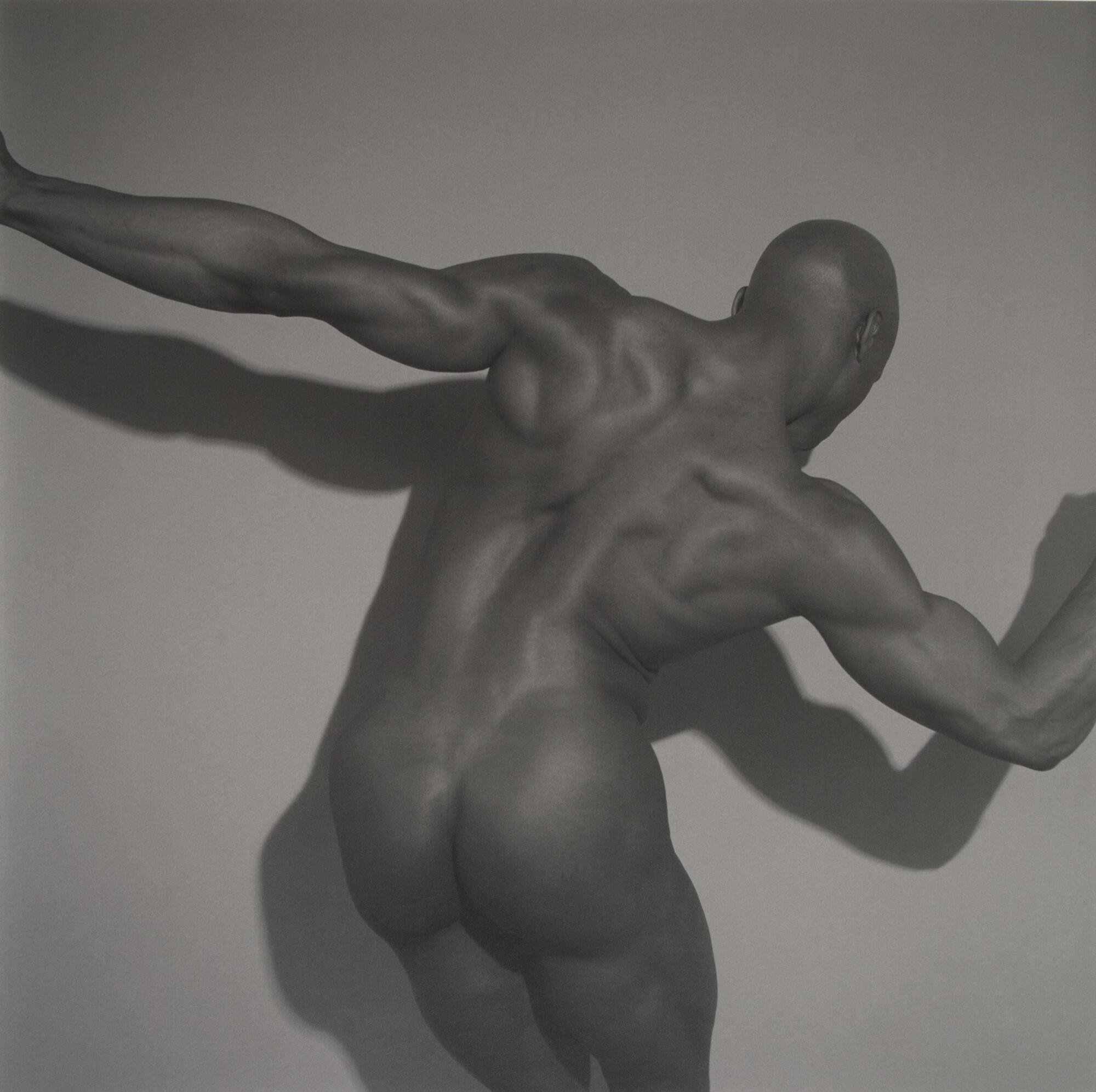 A nude posing in front of a white background. The man is posed with a slight lean and flexing his muscles, with a light source from the top-right casting a shadow on the background wall.