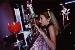 A photograph of a young girl ordering a drink at a bar. She holds her arm up, revealing a watch, and wears sunglasses on her head. A large red drink sits in the foreground on the bar and a neon sign hangs in the background.
