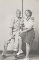 A portrait of a seated couple against a wall with a single stripe in the center of the composition. The man, sitting on the left, wears a military uniform and has his arm around the woman's shoulder. 
