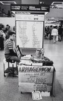 Man seated at a table with Armageddon sign. He is wearing a striped shirt and looking at the camera. There is a New York City bus map posted on the column behind him.