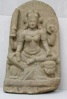 The four-armed Durga sits on a stylized crouching lion with her right leg pendant and the left one across her body.  Her front right hand extends down with palm outwards in a boon giving gesture while the back right hand holds a sword.  Her left font hand holds a fruit or flower bud while her back left hand holds a shield. The whole is simply carved with rather subdued jewelry, but she does wear necklaces, bracelets, armlets and loose anklets as well as large circular earrings and a diadem across her forehead.  Her hair is arranged behind the diadem. The stele is subtly pointed and its only decoration is a band along the outside, although a throne is suggested at her knees.  A highly stylized lotus supports her right foot at the base.<br />